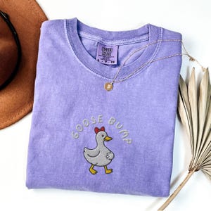 May include: A lavender Comfort Colors t-shirt with the embroidered phrase "GOOSE BUMP" and a cartoon goose wearing a red bow. A gold necklace with a pendant rests on the shirt. A brown hat and dried plant are in the background.