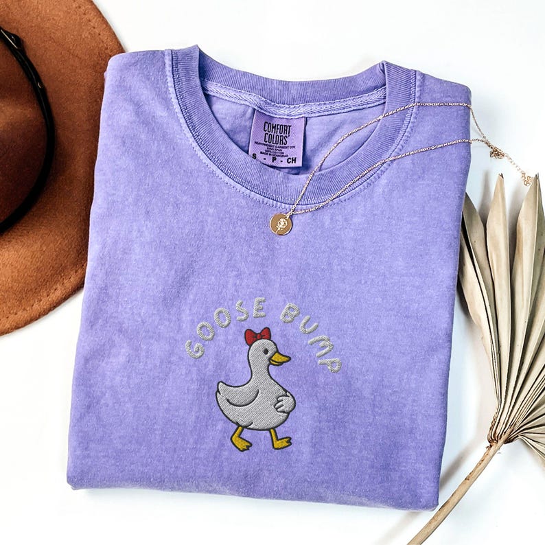 May include: A lavender Comfort Colors t-shirt with the embroidered phrase "GOOSE BUMP" and a cartoon goose wearing a red bow. A gold necklace with a pendant rests on the shirt. A brown hat and dried plant are in the background.