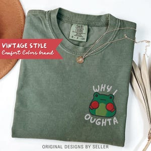 May include: A sage green Comfort Colors brand t-shirt with an embroidered frog design and the words "WHY I OUGHTA". A gold necklace with a pendant rests on the shirt. The words "VINTAGE STYLE" and "ORIGINAL DESIGNS BY SELLER" are also visible.