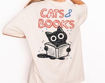 May include: Ivory t-shirt featuring the text "CATS & BOOKS" in red and blue. A black cat reading a book is illustrated below the text. The shirt also has the text "ORIGINAL DESIGNS BY SELLER".