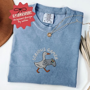 May include: A light blue t-shirt with the words "SERIOUS GAMER" embroidered around a cartoon goose holding a video game controller. The shirt has a small tag that says "COMFORT COLORS". A gold necklace is draped across the shirt.