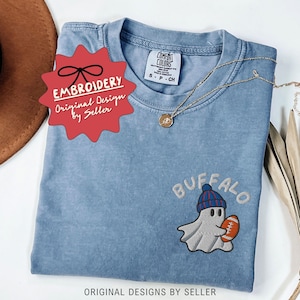 May include: A light blue Comfort Colors t-shirt with an embroidered design of a ghost wearing a blue and red beanie and holding a football. The word "BUFFALO" is embroidered above the ghost. The shirt has a gold necklace and a red tag that says "EMBROIDERY Original Design by Seller".