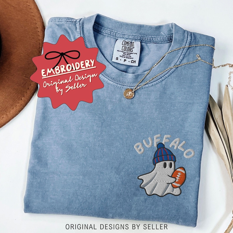 May include: A light blue Comfort Colors t-shirt with an embroidered design of a ghost wearing a blue and red beanie and holding a football. The word "BUFFALO" is embroidered above the ghost. The shirt has a gold necklace and a red tag that says "EMBROIDERY Original Design by Seller".