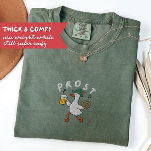 May include: A sage green t-shirt with the word "PROST" embroidered on the front, along with a cartoon goose holding a beer and pretzel. The shirt has the text "THICK & COMFY nice weight while still super comfy" on a red banner. A gold necklace is draped on the shirt.