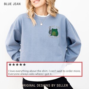 May include: Blue jean-colored sweatshirt with a green frog graphic and the text "WHAT THE FUCK, OVER." The sweatshirt is a light blue color and has a crew neck. The design is embroidered on the front.