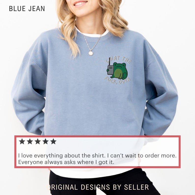 May include: Blue jean-colored sweatshirt with a green frog graphic and the text "WHAT THE FUCK, OVER." The sweatshirt is a light blue color and has a crew neck. The design is embroidered on the front.