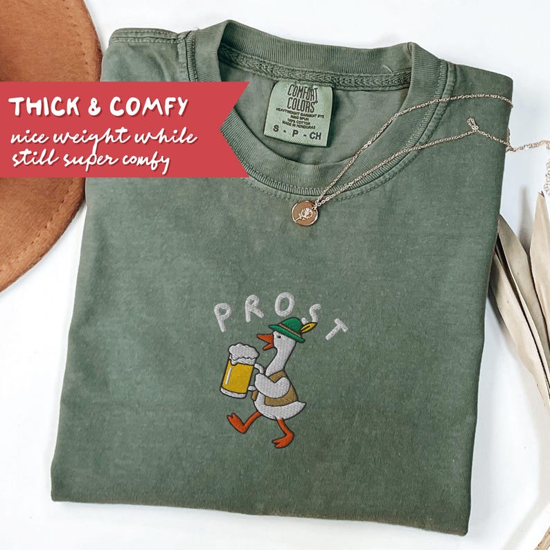 May include: Green Comfort Colors t-shirt with the word "PROST" embroidered above a cartoon goose holding a beer stein. The shirt has the text "THICK & COMFY nice weight while still super comfy" on a red banner. A silver necklace is draped on the shirt.