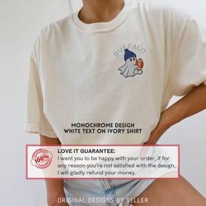 May include: Ivory-colored t-shirt featuring an embroidered ghost wearing a blue beanie and holding a football. The word "Buffalo" is arched above the ghost. The shirt has the text "Monochrome Design White Text on Ivory Shirt".
