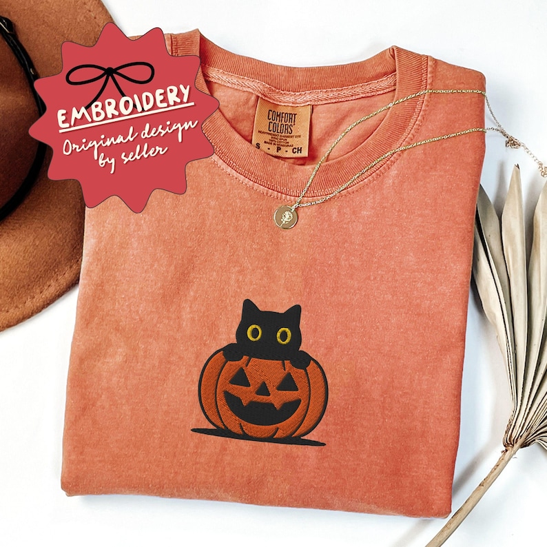 K&ouml;nnte beinhalten: Ein orangefarbenes Comfort Colors T-Shirt mit einer schwarzen Katze, die aus einem geschnitzten K&uuml;rbis lugt. Das Shirt hat eine goldene Halskette und ein Etikett mit der Aufschrift "Comfort Colors". Ein rotes Etikett mit "EMBROIDERY" befindet sich oben links.