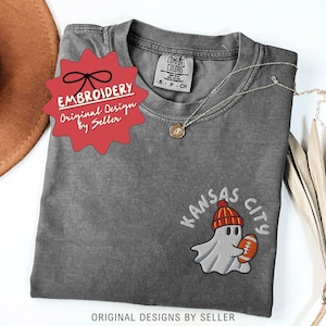 May include: A gray Comfort Colors t-shirt with an embroidered design. The design features a ghost wearing a red and yellow beanie, holding a football, with "Kansas City" above it. The shirt has a small tag that says "S-P-CH".