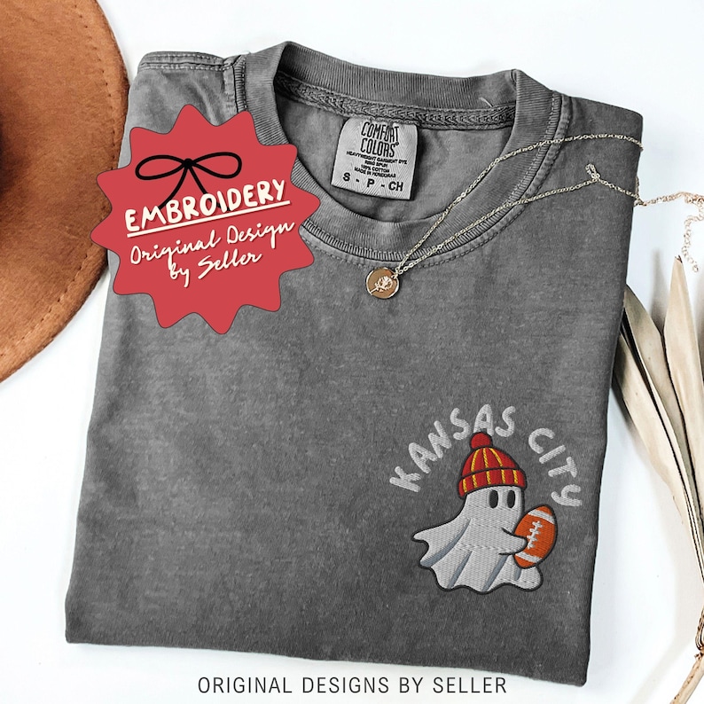 May include: A gray Comfort Colors t-shirt with an embroidered design. The design features a ghost wearing a red and yellow beanie, holding a football, with "Kansas City" above it. The shirt has a small tag that says "S-P-CH".