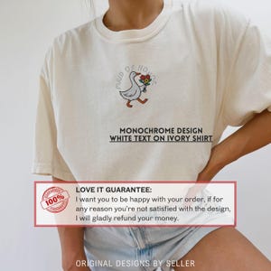 May include: Ivory-colored t-shirt featuring an embroidered cartoon goose carrying flowers, with the text "MAID OF HONOR." Below the goose is the text "MONOCHROME DESIGN WHITE TEXT ON IVORY SHIRT."