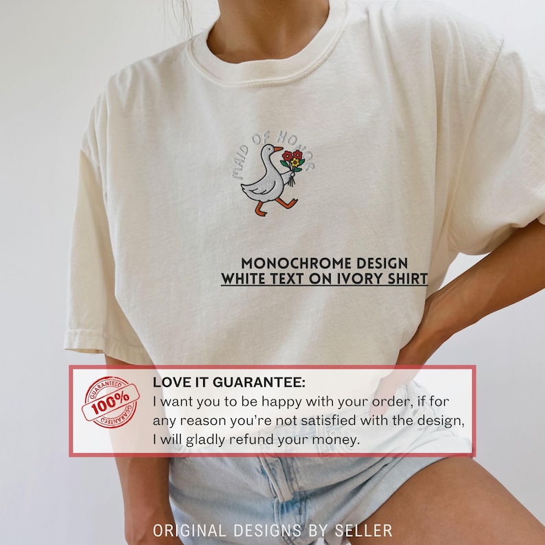 May include: Ivory-colored t-shirt featuring an embroidered cartoon goose carrying flowers, with the text "MAID OF HONOR." Below the goose is the text "MONOCHROME DESIGN WHITE TEXT ON IVORY SHIRT."