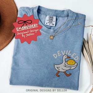May include: A blue Comfort Colors t-shirt with an embroidered design of a goose carrying a deviled egg. The word "DEVILED" is embroidered above the goose. A gold necklace is draped across the shirt. The shirt has a tag that says "S-P-CH".