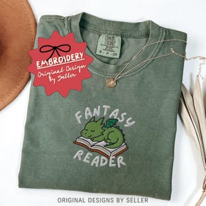 May include: Olive green t-shirt with embroidered design. The design features a green dragon sleeping on an open book, with the words "FANTASY READER" embroidered below. The shirt also has the text "EMBROIDERY Original Design by Seller". A gold necklace is draped across the shirt.