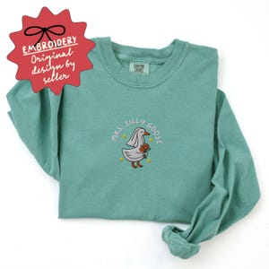 May include: A long-sleeved, teal-colored shirt with an embroidered design. The design features a cartoon goose wearing a veil and holding flowers, with the text "Mrs. Silly Goose" around it. The shirt has a tag that says "Comfort Colors".