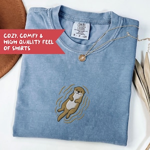 May include: A light blue t-shirt with an embroidered otter design. The shirt has a crew neck and short sleeves. The text "COZY, COMFY & HIGH QUALITY FEEL OF SHIRTS" is in a red banner. A gold necklace with a pendant is on the shirt.