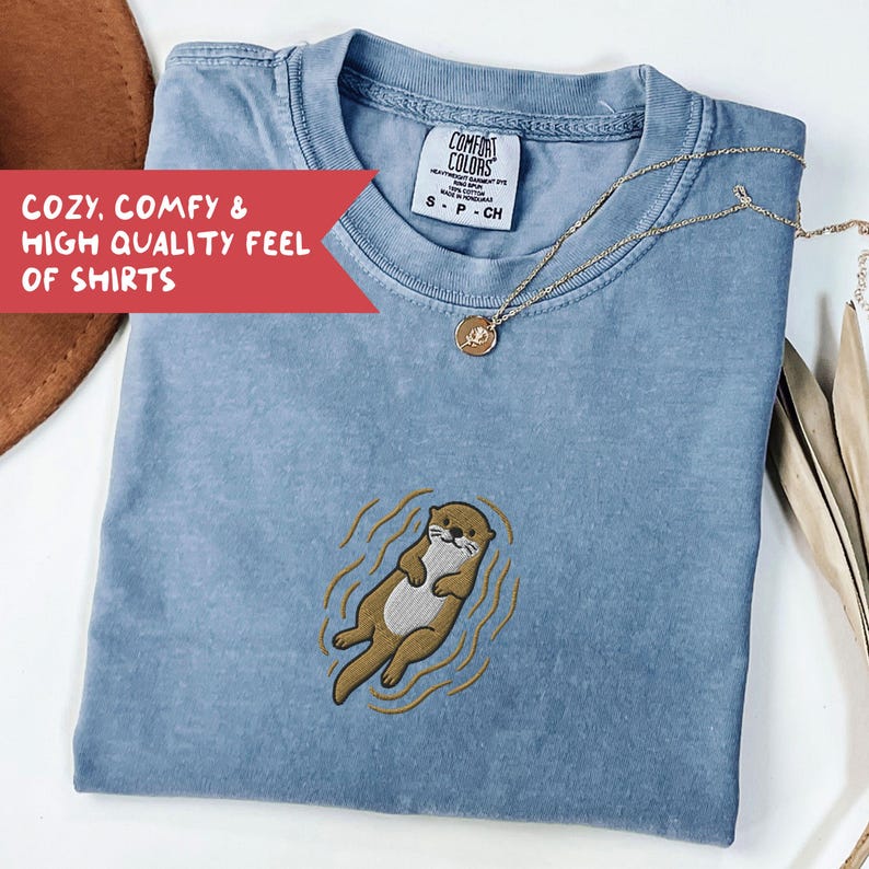May include: A light blue t-shirt with an embroidered otter design. The shirt has a crew neck and short sleeves. The text "COZY, COMFY & HIGH QUALITY FEEL OF SHIRTS" is in a red banner. A gold necklace with a pendant is on the shirt.