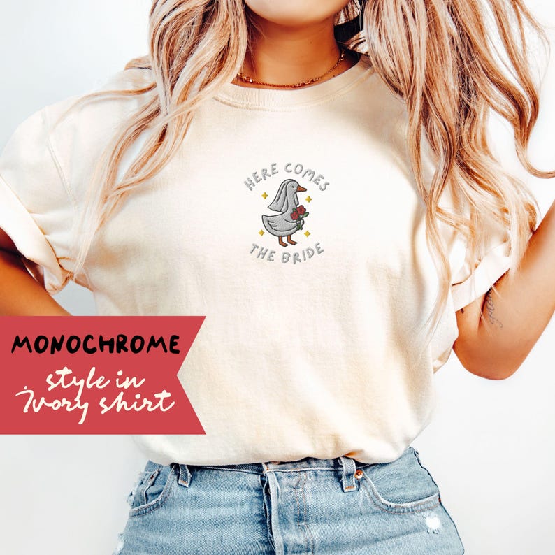 May include: Ivory-colored t-shirt featuring a cartoon goose wearing a veil and holding flowers, with the text "Here Comes The Bride." The shirt has a monochrome style and is made of soft material.
