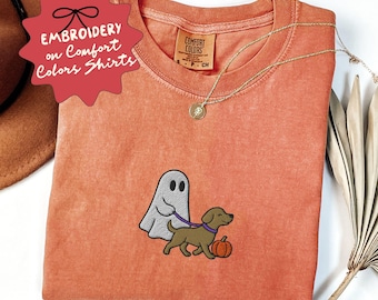 May include: A coral-colored Comfort Colors t-shirt with an embroidered design of a ghost walking a dog on a leash next to a pumpkin. The shirt has a gold necklace and the text "EMBROIDERY on Comfort Colors Shirts" on a red tag.