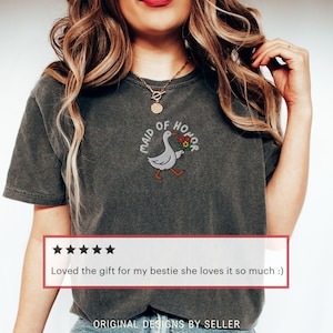 May include: Dark gray t-shirt with an embroidered design of a goose carrying flowers, with the text "MAID OF HONOR". The shirt also has a customer review that says "Loved the gift for my bestie she loves it so much :)".