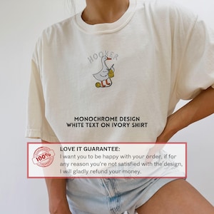 May include: Ivory-colored t-shirt with a humorous embroidered design. The design features a cartoon goose holding a crochet hook and yarn, with the word "HOOKER" above. Below the design, the text reads "MONOCHROME DESIGN WHITE TEXT ON IVORY SHIRT."
