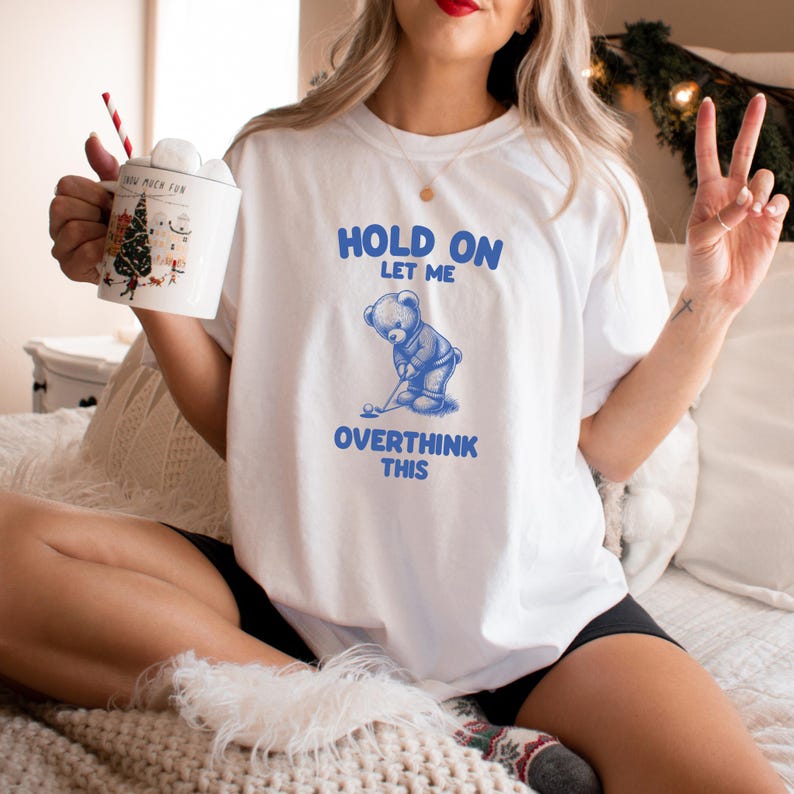 K&ouml;nnte beinhalten: Wei&szlig;es T-Shirt mit blauem Text, der "Hold on let me overthink this" lautet. Ein blauer Comic-B&auml;r h&auml;lt einen Golfschl&auml;ger und ein Golfball liegt auf dem Boden.