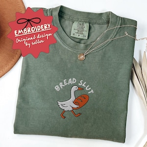 May include: Olive green t-shirt with the embroidered phrase "BREAD SLUT" and a cartoon goose carrying a loaf of bread. The shirt has a Comfort Colors tag and a small gold necklace. A red starburst tag reads "EMBROIDERY Original design by seller."