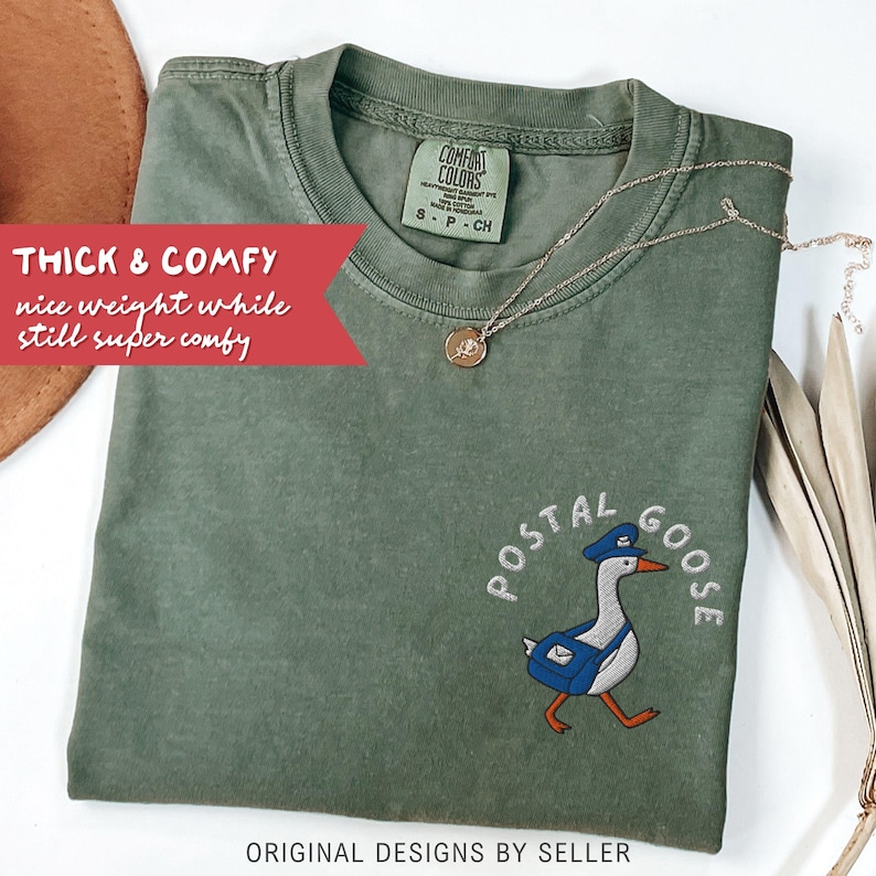 May include: An olive green t-shirt with the embroidered image of a goose wearing a postal worker hat and carrying a mail bag. The text "POSTAL GOOSE" is above the goose. A gold necklace with a pendant is on the shirt.