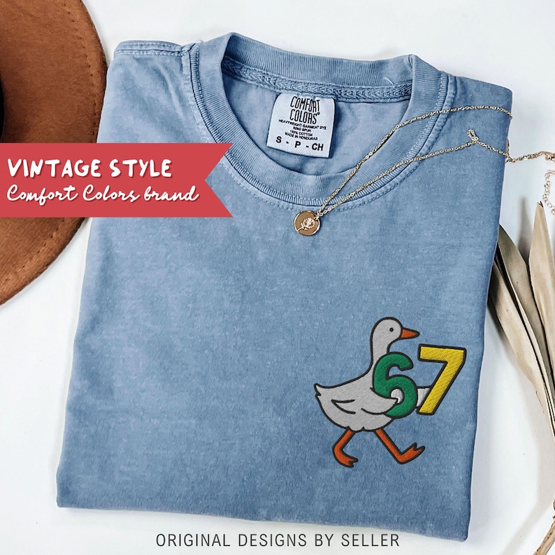 May include: A light blue Comfort Colors brand t-shirt with a cartoon duck carrying the number 67. The shirt has a vintage style and the text "ORIGINAL DESIGNS BY SELLER". A gold necklace is draped across the shirt.