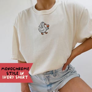 May include: Cream-coloured t-shirt featuring an embroidered cartoon goose holding a wine glass and wearing a veil. The shirt has a crew neck and short sleeves. The text "MONOCHROME STYLE IN IVORY SHIRT" is on a red banner.