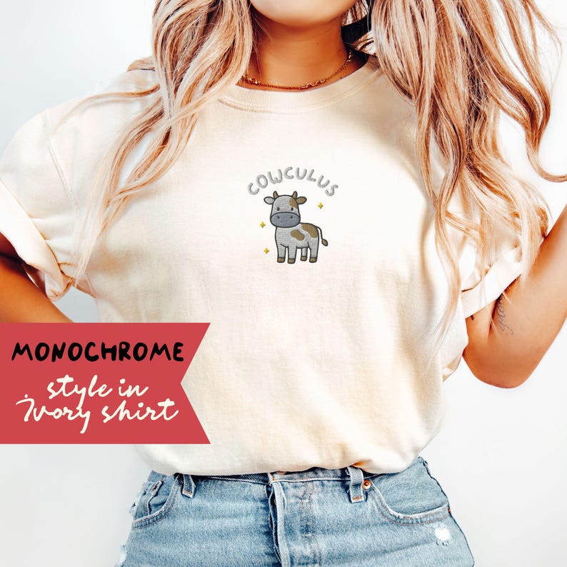 May include: Ivory-colored t-shirt featuring a cartoon cow illustration with the word "COWCULUS" above it. The shirt has a relaxed fit and is made of a soft material. The text "MONOCHROME style in Ivory shirt" is printed on a red banner.