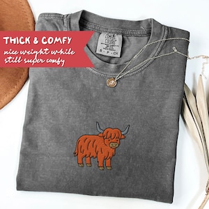May include: A gray Comfort Colors t-shirt with an embroidered orange Highland cow design. The shirt has a crew neck and short sleeves. Text on the shirt reads "THICK & COMFY nice weight while still super comfy". A silver necklace with a pendant is also visible.