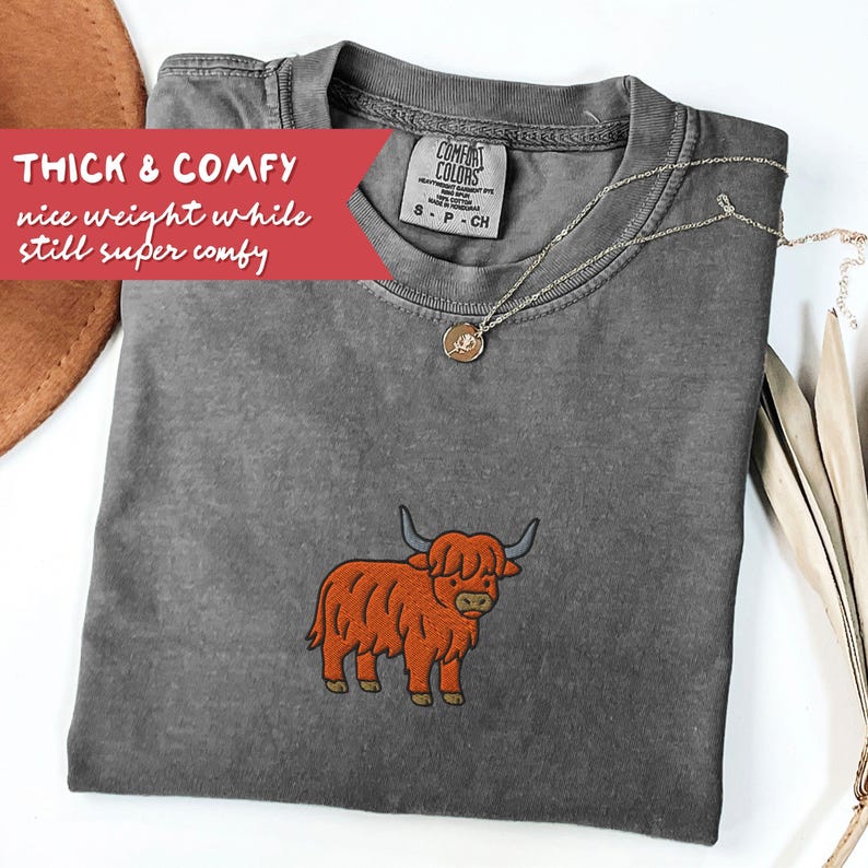 May include: A gray Comfort Colors t-shirt with an embroidered orange Highland cow design. The shirt has a crew neck and short sleeves. Text on the shirt reads "THICK & COMFY nice weight while still super comfy". A silver necklace with a pendant is also visible.