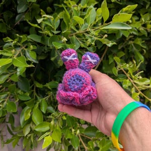 May include: A small, round, crocheted plush toy featuring a variegated purple and pink yarn pattern. The object has two upright, rounded ears with white stitched X-shaped details on each, and a ruffled base made of the same purple and pink yarn.