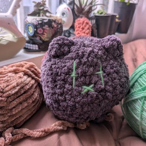 May include: A dark purple, round, crocheted plush toy shaped like a cat head with small ears. The face features embroidered light green thread forming two vertical lines for eyes and a small cross for a mouth. The texture is soft and fuzzy.