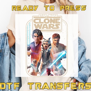 May include: Cream-colored t-shirt featuring a graphic from Star Wars: The Clone Wars. The design includes characters with lightsabers and the text "READY TO PRESS" and "DTF TRANSFERS" in a gold font.