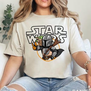 May include: A beige t-shirt with a cartoon image of a Mandalorian and Grogu. The text "STAR WARS" is printed in black above the image.