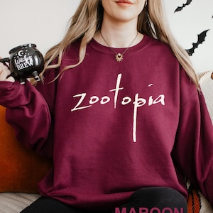 May include: A maroon sweatshirt with the word "Zootopia" in white script. The word "MAROON" is printed at the bottom of the image. A black mug with the words "Witches Brew" is held in the left hand.