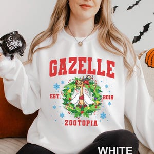 Op de afbeelding: Witte sweatshirt met het woord "GAZELLE" in rode blokletters. Daaronder een krans met een cartoon gazelle met een rood topje. De tekst "EST. 2016" en "ZOOTOPIA" staan ook op de sweatshirt. Het woord "WHITE" staat onderaan.