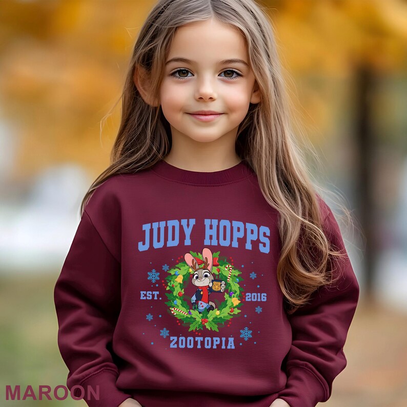 Op de afbeelding: Bordeaux sweatshirt met de tekst "JUDY HOPPS" in blauw, en een cartoonafbeelding van Judy Hopps in een krans. De sweatshirt heeft ook de tekst "EST. 2016" en "ZOOTOPIA" in blauw. Het woord "MAROON" staat onderaan.