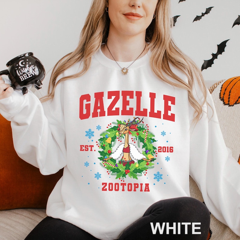 Op de afbeelding: Witte sweatshirt met het woord "GAZELLE" in rode blokletters. Daaronder een krans met een cartoon gazelle met een rood topje. De tekst "EST. 2016" en "ZOOTOPIA" staan ook op de sweatshirt. Het woord "WHITE" staat onderaan.
