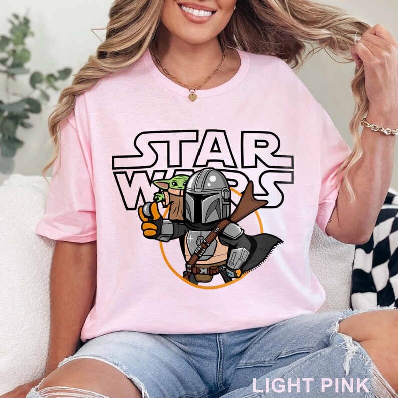 May include: A light pink t-shirt with a cartoon image of a Mandalorian and Grogu from Star Wars. The text "STAR WARS" is printed in black above the image.