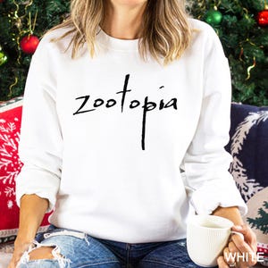 May include: A white sweatshirt featuring the word "Zootopia" in black cursive lettering. The person is holding a white mug. The background includes a Christmas tree and decorative cushions.