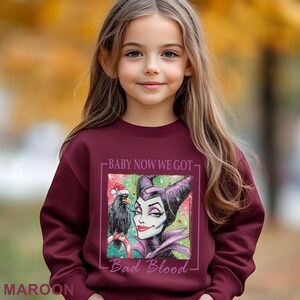 Puede incluir: Una sudadera granate con un gr&aacute;fico de Mal&eacute;fica y un cuervo con un gorro de Pap&aacute; Noel. El texto en la sudadera dice "BABY NOW WE GOT - Bad Blood". La sudadera la lleva una ni&ntilde;a.