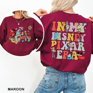 May include: Maroon sweatshirt featuring a colorful graphic of Disney and Pixar characters on the front and back. The back of the sweatshirt has the text "IN MY DISNEY PIXAR ERA" in a retro font. The front has a collage of characters.
