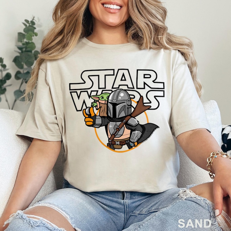 May include: A beige t-shirt with a cartoon image of a Mandalorian and Grogu. The text "STAR WARS" is printed in black above the image.