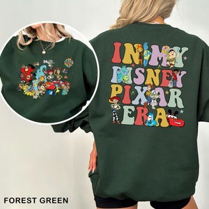 May include: Forest green sweatshirt featuring a colorful graphic of Disney Pixar characters on the front and the phrase "IN MY DISNEY PIXAR ERA" on the back. The sweatshirt has a crew neck and long sleeves, perfect for casual wear.