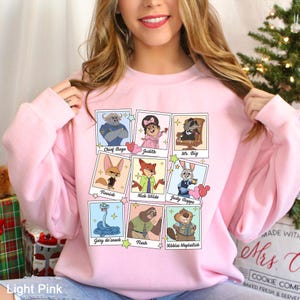 Puede incluir: Sudadera rosa claro con una cuadr&iacute;cula de retratos de personajes de dibujos animados. Los personajes incluyen a Chief Bogo, Judith, Mr. Big, Finnick, Nick Wilde, Judy Hopps, Gary de'snack, Flash y Nibbles Maplestick. El texto "Light Pink" est&aacute; en la parte inferior.
