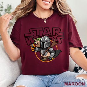 May include: A maroon t-shirt with a graphic of a cartoon Mandalorian and Grogu from Star Wars. The text "STAR WARS" is printed above the characters.